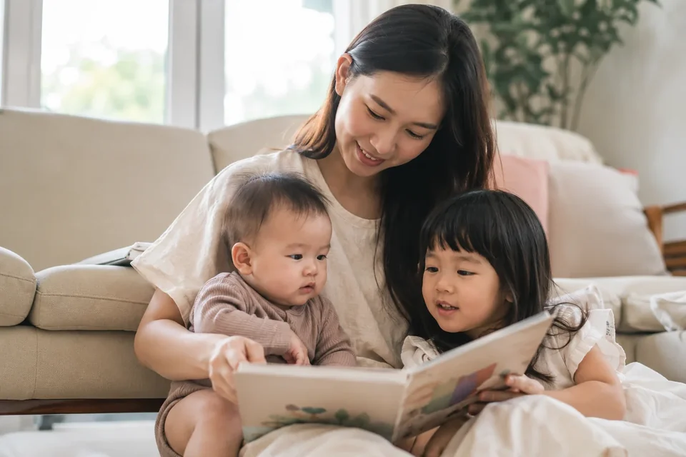 คุณแม่อุ้มทารกและอ่านหนังสือกับพี่สาววัยอนุบาลในห้องนั่งเล่น